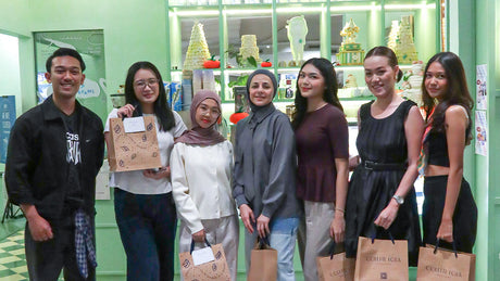 A group of seven people standing together inside Colette Lola Café Studio, smiling and holding Colette Lola gift bags in front of a display of cakes and decorations
